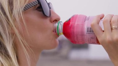 Genç, beyaz, sarışın bir kadın açık havada boynundan taze limonata içiyor. Meyve suyu şişesiyle güzel bir kızın, limonatayla poz veren sağlıklı bir kadının yakın çekimi. Detokslu diyet yemeği.