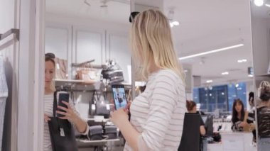 Alışveriş konsepti, moda, tarz, teknoloji ve insanlar - akıllı telefonlu mutlu kadın aynada aynada selfie çekiyor giyim mağazasında çanta ya da moda kıyafetleri konsepti deneyen müşteri seçiyor..