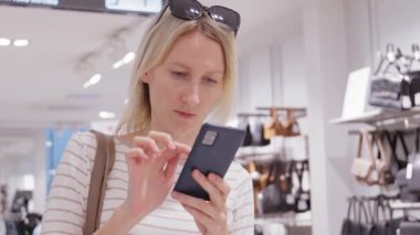 Giyim mağazasından alışveriş yapan güzel bir bayan, akıllı telefon kullanıyor, internette geziniyor, internetten karşılaştırıyor, şık kıyafetler seçiyor. Moda mağazası, renkli markalar.,