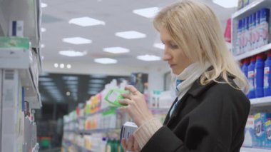 Kadın kozmetik ürünlerini dükkanda seçiyor. Siyah paltolu güzel sarışın kadın elinde bir kutu tutuyordu. Yandan bak. Kişisel bakım ürünleri ve modern kozmetoloji.