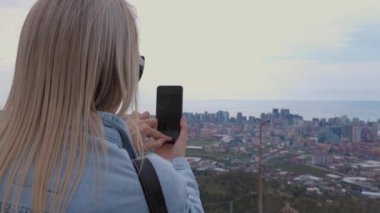 Turistlerin elinde akıllı telefon, Batumi City, Georgia 'nın fotoğrafını çekiyor. Kot ceketli sarışın kız deniz kıyısında ftografçı şehir manzaralı. .