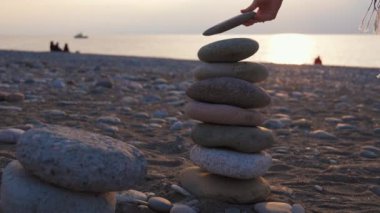 Ruhsal olarak rahat bir kadın gün batımında çakıl taşlı kumsalda kaldırım taşlarının piramidine taş koyuyor. Yoga kadın iç huzuru rahatlat meditasyon uyumu huzur dengesi dengesi piramidi
