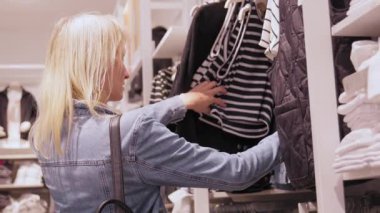 Moda mağazasında durup kıyafet seçen mutlu bir kadın. Şık giysi raflarını inceliyor, karar vermeden önce her birini dikkatlice inceliyor..