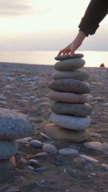 Ruhsal olarak rahat bir kadın gün batımında çakıl taşlı kumsalda kaldırım taşlarının piramidine taş koyuyor. Yoga kadın iç huzuru rahatlat meditasyon uyumu huzur dengesi dengesi piramidi