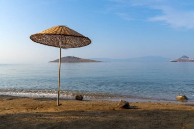 Kumlu bir plajın kıyısında örülmüş bir plaj şemsiyesinin yakın çekimi sabahın erken saatlerinde denizde küçük bir adanın arka planına karşı. Güzel deniz turizmi manzarası