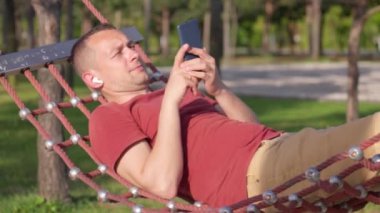 Kulaklıklı genç adam tropik bahçede tatilde dinleniyor. - Hamaklı, kulaklıklı bir adam. Yaz parkında ya da bahçede telefondan gelen ses kayıtlarını dinliyor. Mutlu Adam Yaz Tatili