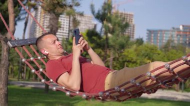 Kafkasyalı genç adam akıllı telefon kullanıyor, kulaklıkla müzik dinliyor, bahçedeki hamakta, rahatlatıcı ve yaz konseptini internet ya da sosyal medya kullanarak ve mobil uygulamaları tarıyor..