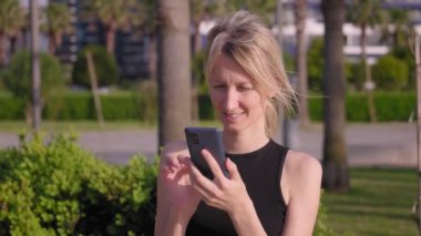 Yaz parkında, kadın nüfuzlu birinin portresi, cep telefonu kullanmak, okumak ve örnek telefon görüntülerine bakmak, açık havada siyah giyinmek, rahatlamak, cep telefonu kullanmak. Kentsel Yaşam Tarzı