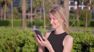 Yaz parkında, kadın nüfuzlu birinin portresi, cep telefonu kullanmak, okumak ve örnek telefon görüntülerine bakmak, açık havada siyah giyinmek, rahatlamak, cep telefonu kullanmak. Kentsel Yaşam Tarzı