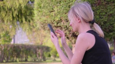 Yaz parkında, kadın nüfuzlu birinin portresi, cep telefonu kullanmak, okumak ve örnek telefon görüntülerine bakmak, açık havada siyah giyinmek, rahatlamak, cep telefonu kullanmak. Kentsel Yaşam Tarzı