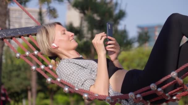 Caucasian Young Blonde Woman Using Smartphone Listening Music Headphones Hammock — ストック動画