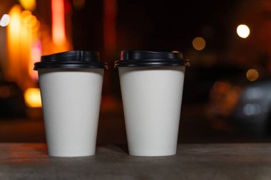 Bulanık bir gece şehrinin arka planında kahve ya da çay eşliğinde iki beyaz çevre dostu fincanın yakın çekimi. Kahveyi yanına al..