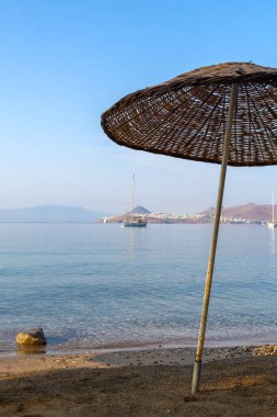 Kumlu bir plajın kıyısındaki örülmüş bir plaj şemsiyesinin yakın çekimi deniz yüzeyinin arka planına, yatlara, sabahın erken saatlerinde evlere karşı. Dikey fotoğraf