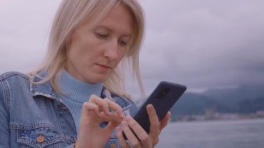 Bulutlara ve dağlara karşı Denim takım elbiseli mutlu beyaz kadın, akıllı telefon kullanıyor, görüntüye bakıyor, ciddi ve gergin, cep telefonu okuyor..