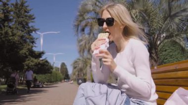 Siyah güneş gözlüklü mutlu sarışın bir kadının portresi. Güneşli yaz gününde parktaki bankta oturmuş hazır shawarma yiyor. Sağlıksız yiyecekler, yemek molası konseptleri. Arka planda palmiye ağaçları