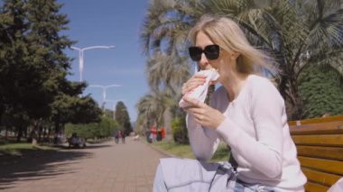 Siyah güneş gözlüklü mutlu sarışın bir kadının portresi. Güneşli yaz gününde parktaki bankta oturmuş hazır shawarma yiyor. Sağlıksız yiyecekler, yemek molası konseptleri. Arka planda palmiye ağaçları