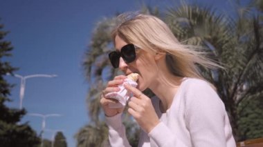 Siyah güneş gözlüklü mutlu sarışın kadının yakın plan portresi. Güneşli yaz gününde parkta bankta oturmuş hazır shawarma yiyor. Sağlıksız yiyecekler, yemek molası konseptleri. Arka planda palmiye ağaçları