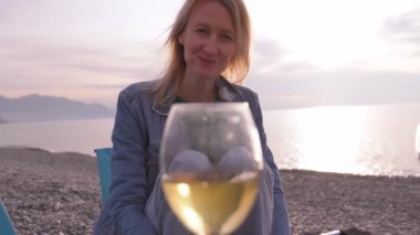 Genç bir kadın deniz kıyısında oturuyor ve lezzetli hamur turtalarını yiyor, ellerinin arasında tutuyor ve yanında iki kadeh şarap var. Gülümser, konuşur ve mutlu bir şekilde gülümser. Piknik denizi Sahilde keyif