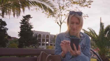 Palmiye ağaçlarıyla parktaki akıllı telefonunu kontrol eden genç bir kadın. Günlük kıyafetli kız akıllı telefon tutuyor, mesaj yazıyor, parkta otururken internetten alışveriş yapıyor..