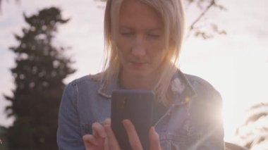 Palmiye ağaçlarıyla parktaki akıllı telefonunu kontrol eden genç bir kadın. Günlük kıyafetli kız akıllı telefon tutuyor, mesaj yazıyor, parkta otururken internetten alışveriş yapıyor..