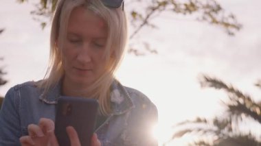 Palmiye ağaçlarıyla parktaki akıllı telefonunu kontrol eden genç bir kadın. Günlük kıyafetli kız akıllı telefon tutuyor, mesaj yazıyor, parkta otururken internetten alışveriş yapıyor..