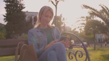 Palmiye ağaçlarıyla parktaki akıllı telefonunu kontrol eden genç bir kadın. Günlük kıyafetli kız akıllı telefon tutuyor, mesaj yazıyor, parkta otururken internetten alışveriş yapıyor..