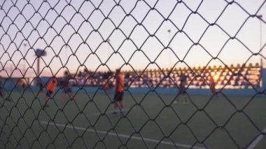 Tanınmayan oyuncular tarafından oynanan futbol, arka planda denizle birlikte gün batımında açık gökyüzünün altında metal çit örgüsü ya da futbol sahasında çekilen. Sahilde Günbatımı Oyun Alanı.