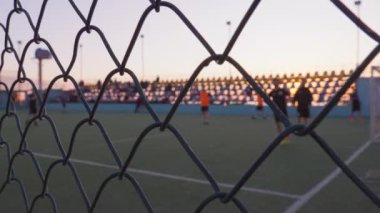 Tanınmayan oyuncular tarafından oynanan futbol, arka planda denizle birlikte gün batımında açık gökyüzünün altında metal çit örgüsü ya da futbol sahasında çekilen. Sahilde Günbatımı Oyun Alanı.
