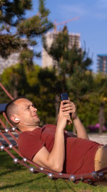 Genç adam dışarıda bir hamakta dinlenirken kablosuz kulaklıkla müzik dinliyor. Mutlu adam parktaki bir hamakta şort ve tuğla renkli tişört giyerek rahatlıyor.