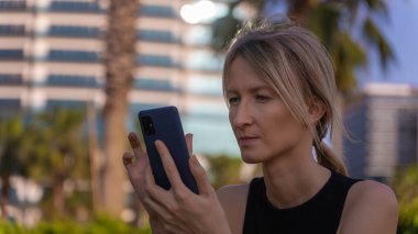 Atkuyruklu, odaklanmış genç sarışın bir kadının parkta cep telefonuna mesaj yazıp, okuduğu yeri kopyaladığı yakın plan. Kadın güneşli bir günde parkta telefonuna bakıyor.