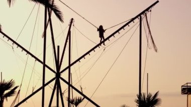Çocuk trambolinde günbatımına doğru yükseğe zıplıyor ve elastik bantlı elastik kayışla güneş. Neşeli küçük kız trambolinde trambolinde, lunaparkta, parlak elastik bantlarda uçuyor.