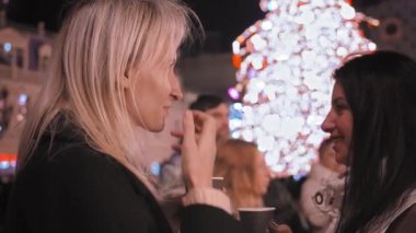 İki beyaz kadın festival meydanında durmuş gülümsüyor, sohbet ediyor ve Noel 'i arka planda kutluyorlar. Kahve iç ya da bardaktan şarap iç..
