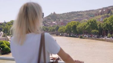 Sarışın, dikiz aynalı, köprüde duran Kura Nehri 'nin Tiflis' ten akan manzarasına hayran. Canlı şehir manzarası ve huzurlu atmosfer mükemmel bir yaz anı yaratır.