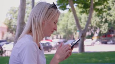 Parkta bankta oturan genç bir kadın, akıllı telefonunu kullanırken gülümsüyor. Açık havada güneşli bir gün geçiriyor, sosyal medyada geziniyor ve internet üzerinden arkadaşlarıyla bağlantı kuruyor.