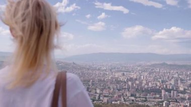Kadın tepenin üstünde duruyor, saçları rüzgarda uçuşuyor, Georgia 'nın genişleyen Tiflis kentinin panoramik manzarasını emiyor. Sahne dinginlik ve ilham anlarını yakalar