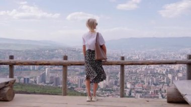 Turist ahşap gözlem güvertesinde duruyor, çitlere yaslanıyor, Gürcistan 'daki Tblisi şehrinin nefes kesici manzarasının keyfini çıkarıyor. Canlı şehir manzarası rahatlamak için mükemmel bir ortam yaratır.