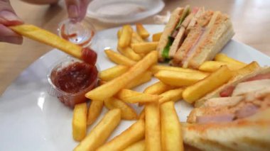 Restoran masasında oturan müşteri patates kızartmasını ketçabın içine daldırıyor. Tabakta kızarmış patates kızartmasının yanında kulüp sandviçi var.