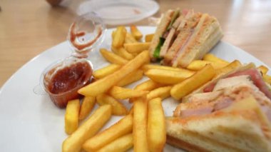 Restoran masasında oturan müşteri patates kızartmasını ketçabın içine daldırıyor. Tabakta kızarmış patates kızartmasının yanında kulüp sandviçi var.