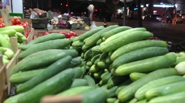 Salatalıklar ve çeşitli sebzeler gece boyunca hareketli sokak pazarında satışa sunuluyor. Arabalar geçiyor ve şehir ışıkları canlı kentsel atmosfer yaratıyor.