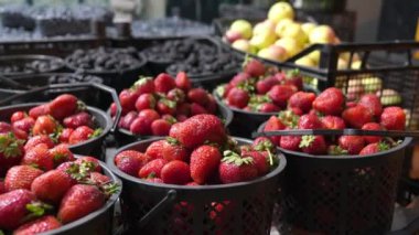 Çiftçi pazarında sepetler dolusu olgun, kırmızı çilek dolup taşıyor. Çilekler üst üste yığılır ve diğer taze ürünlerle çevrilidir.