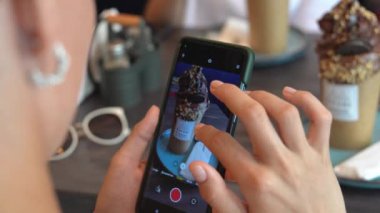 Kadın telefonundaki çikolatalı milkshake fotoğrafının odak noktasını ve çerçevesini dikkatlice ayarlıyor. Milkshake, krem şanti ve çikolata soslu uzun bir bardakta.