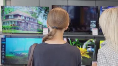 İki kadın, potansiyel müşteri ve satış asistanı, elektronik mağazasında yeni televizyon özelliklerini tartışıyor