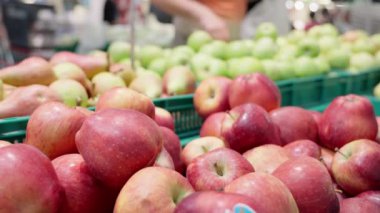 Kırmızı elmalar süpermarkette güzel bir şekilde düzenlenirken, müşteriler arka planda yeşil elmaları seçiyor ve canlı bir market alışverişi ve taze üretim seçenekleri yaratıyorlar.