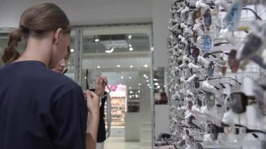 Optik mağazasında güneş gözlüğü seçen iki kadın, farklı modeller deniyorlar. Parlak ekran çeşitli stiller sergiliyor, günlük yaz ortamında alışveriş deneyimini artırıyor