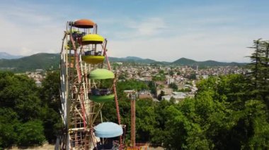 Bereketli yeşil ağaçların arasında dönen parlak dönme dolabın insansız hava aracı görüntüleri güneşli bir günde şehrin ve uzak tepelerin çarpıcı manzarasını sunuyor.