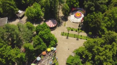 Renkli dönme dolap, güneşli yaz gününde yeşil ağaçlarla çevrili boş lunaparkta yavaşça dönüyor. Manzara nostalji ve dingin yalnızlığı çağrıştırıyor. Hava görüntüsü, İHA görünümü.