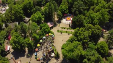 Renkli dönme dolap, güneşli yaz gününde yeşil ağaçlarla çevrili boş lunaparkta yavaşça dönüyor. Manzara nostalji ve dingin yalnızlığı çağrıştırıyor. Hava görüntüsü, İHA görünümü.