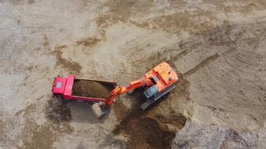 Kazıcı, nehrin yakınındaki inşaat alanında, çöp kamyonuna kum yüklüyor. İnşaat alanında kazıcının çöp kamyonunu yüklerken en tepeden aşağı bakışı. Kazıcı yerde çukur kazıyor.