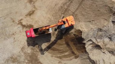 Kazıcı, nehrin yakınındaki inşaat alanında, çöp kamyonuna kum yüklüyor. İnşaat alanında kazıcının çöp kamyonunu yüklerken en tepeden aşağı bakışı. Kazıcı yerde çukur kazıyor.