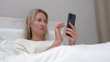 Beyaz çarşaflarla yatakta uzanan kadın akıllı telefonu tutuyor, ekranı tıkırdatıyor ve çeşitli içerikleri internette tarıyor. Rahatlama ve rahatlama sahneyi tanımlar